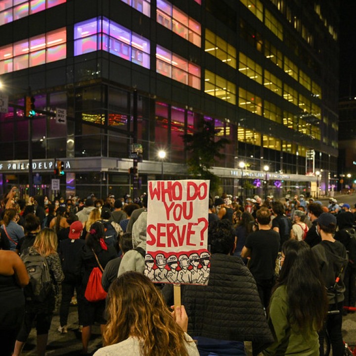 Crowd marches at protest with sign that reads "who do you serve?"