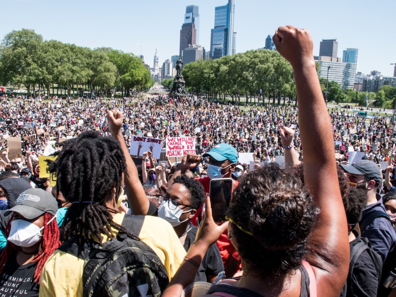 A rally of thousands of people assemble. The philly skyline is in the background and a Black person raises their fist in the foreground.