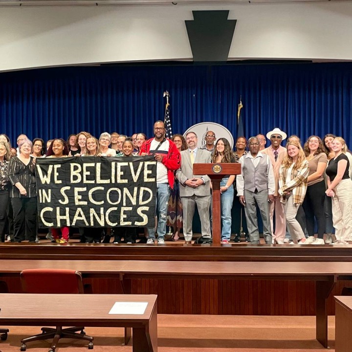A couple dozen people stand on a stage with a banner that reads "we believe in second chances"